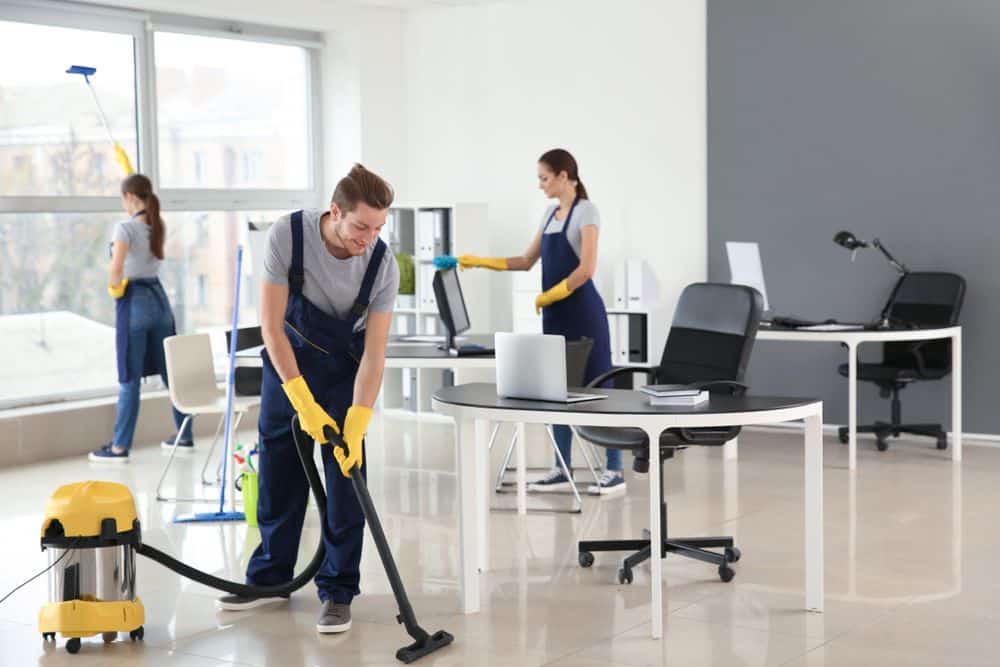 An image of a a group of people, using a cleaning solution and a cloth to wipe down a countertop in a commercial setting. In the background, there are cleaning supplies and equipment such as a vacuum cleaner, mop, and broom, suggesting a thorough and professional commercial cleaning service